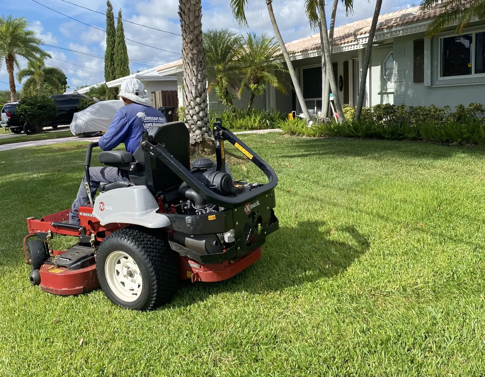 A person is operating a red and white Exmark zero-turn mower to cut grass and lawn care in a residential neighborhood. The mower is in the foreground, with a palm tree and a house in the background.