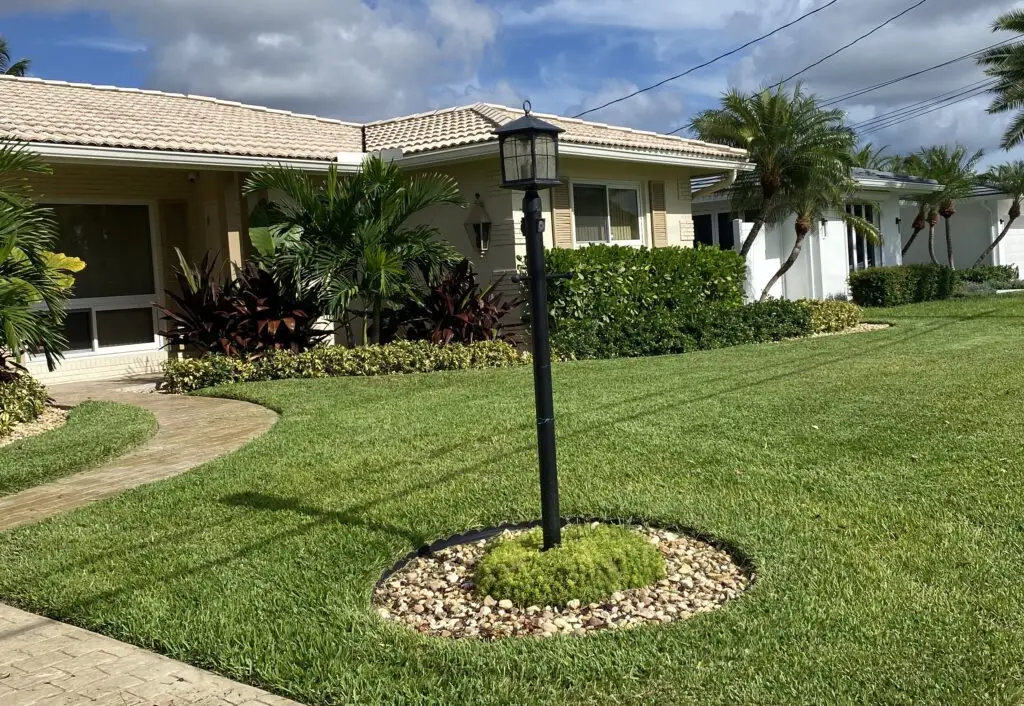 A front yard features a paved walkway, green grass, palm trees, and a lamppost. Two houses with lush lawn care stand in the background.