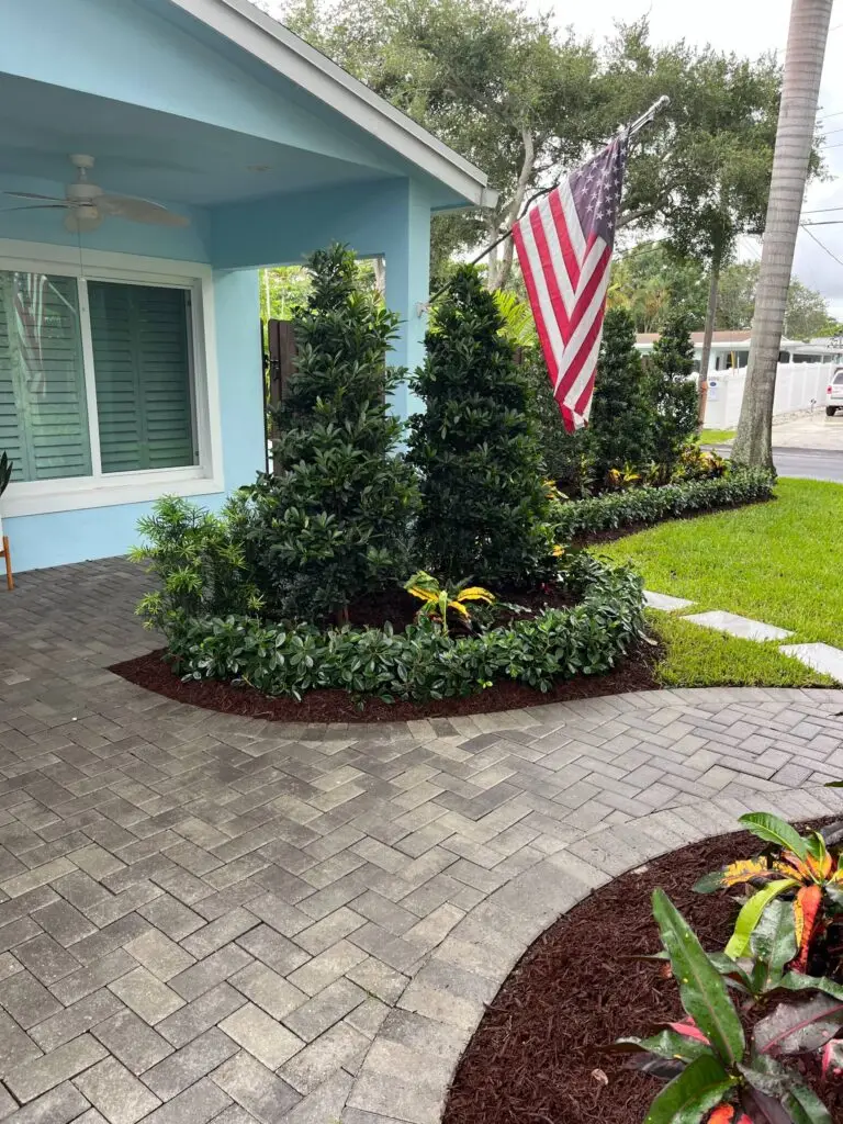 A front yard with a brick walkway, an American flag, and several shrubs and small trees.